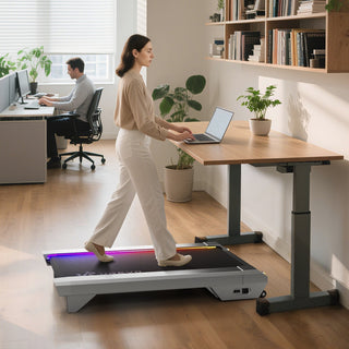 A woman walks on a treadmill desk while using a laptop in a bright office with plants and a colleague working nearby.