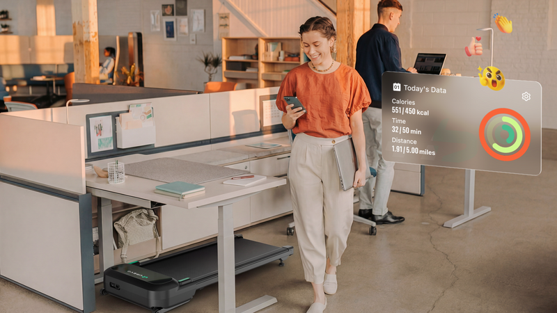 A modern open-plan office with a woman walking beside a desk that has a walking pad treadmill underneath it.