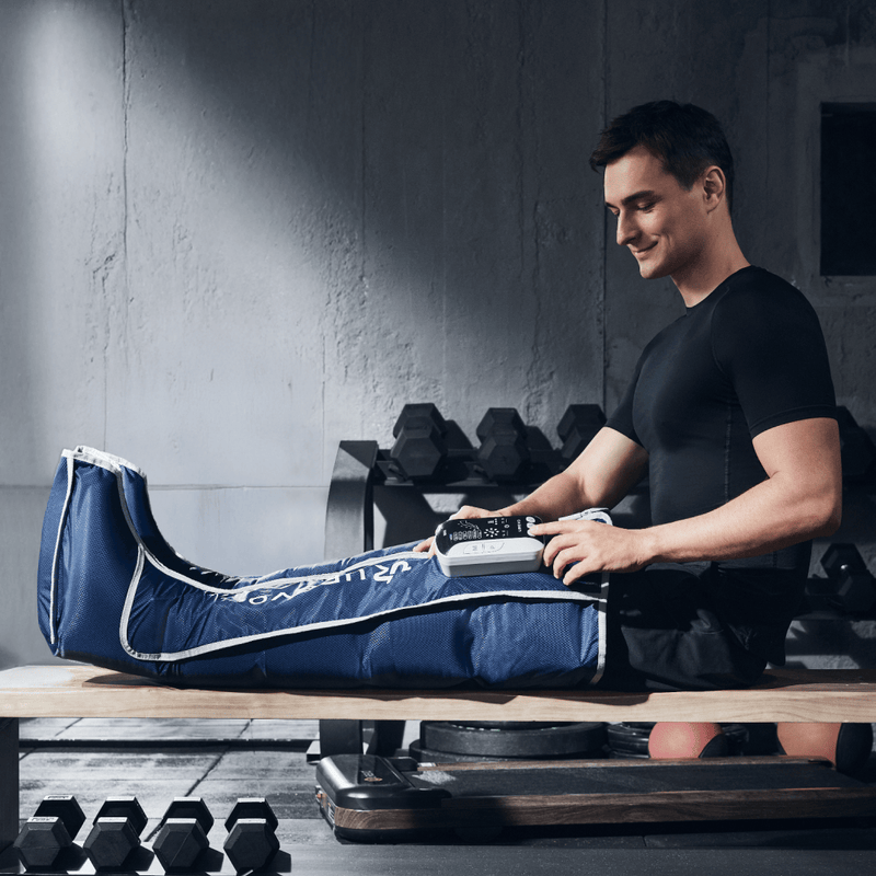 A man in a gym smiles while using a blue leg compression device on his right leg.