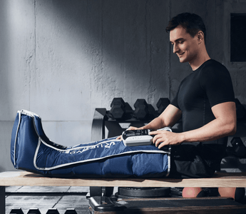 A man in a gym smiles while using a blue leg compression device on his right leg.