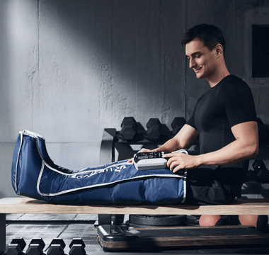A man in a gym smiles while using a blue leg compression device on his right leg.