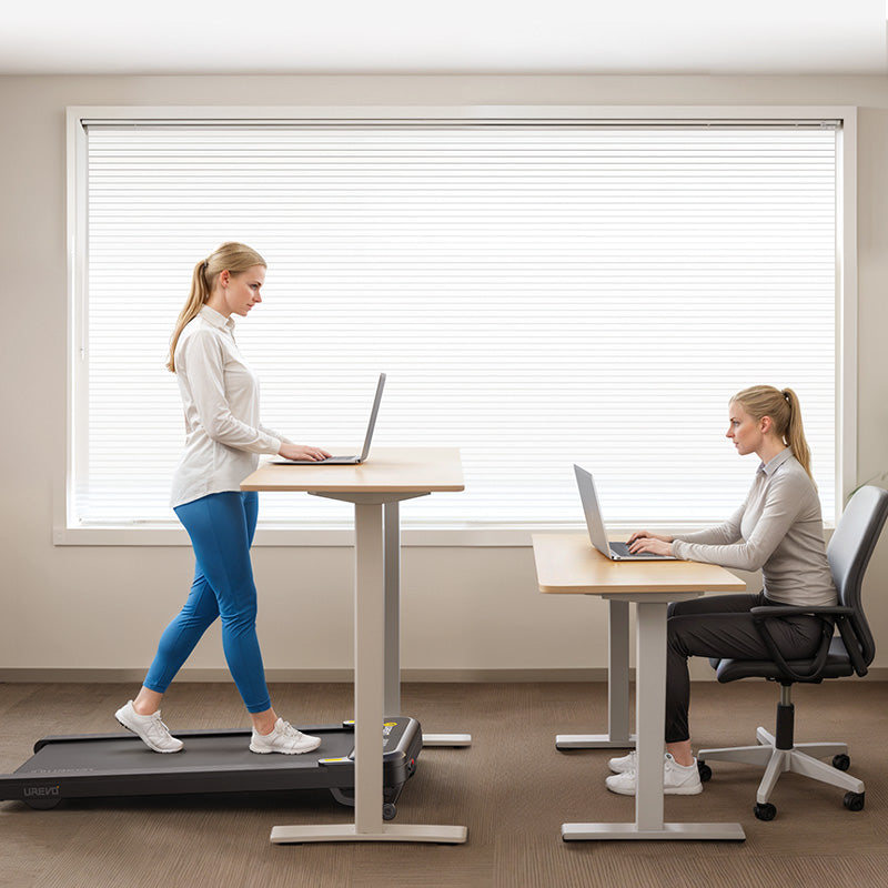 A woman walking on a treadmill desk while using a laptop, with another woman seated at a desk.