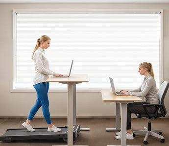 A woman walking on a treadmill desk while using a laptop, with another woman seated at a desk.
